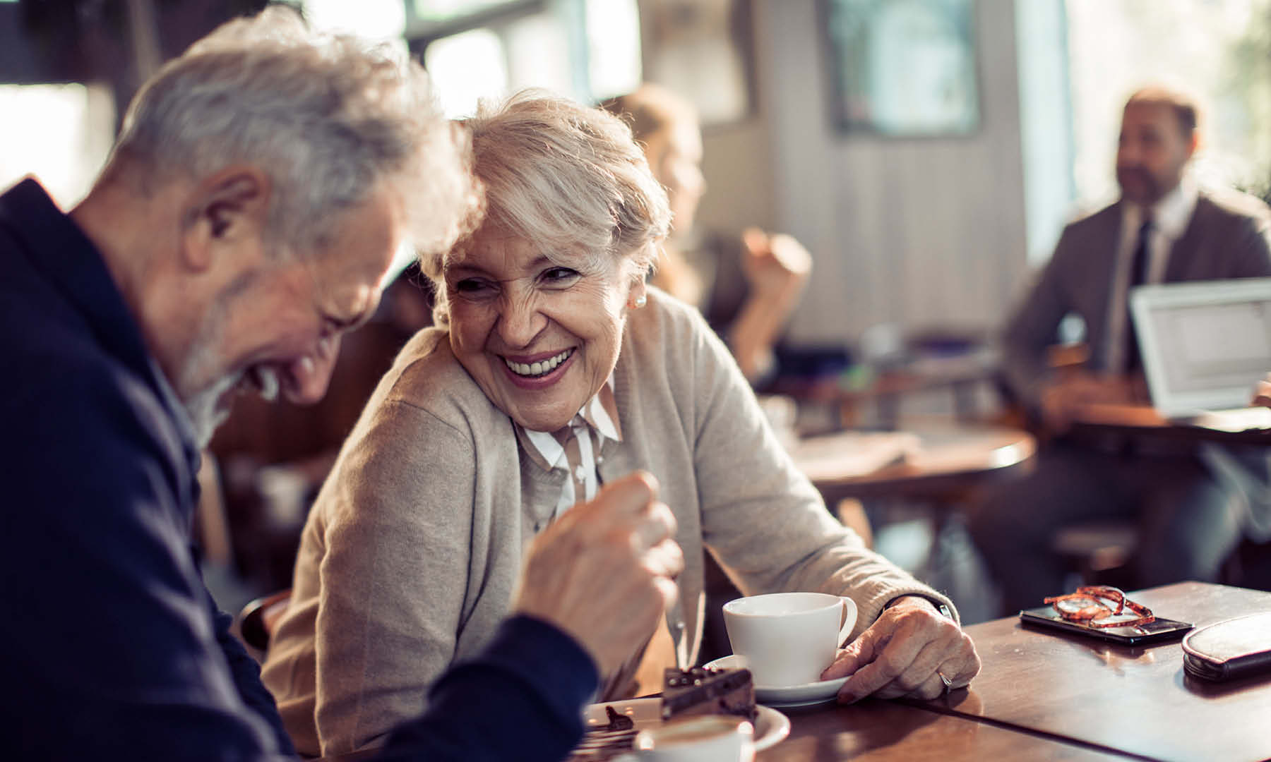 Senior couple laugh and smile sitting at coffee shop close up view with mugs and phone nearby and other coffee shop patrons in background
