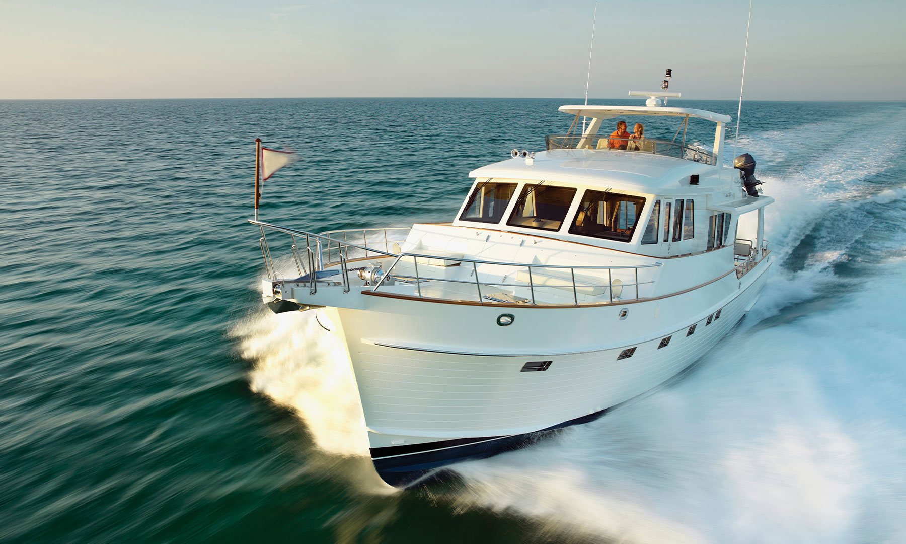 Image of sleek white yacht boat speeding across teal waters at dusk with a few people on the top.