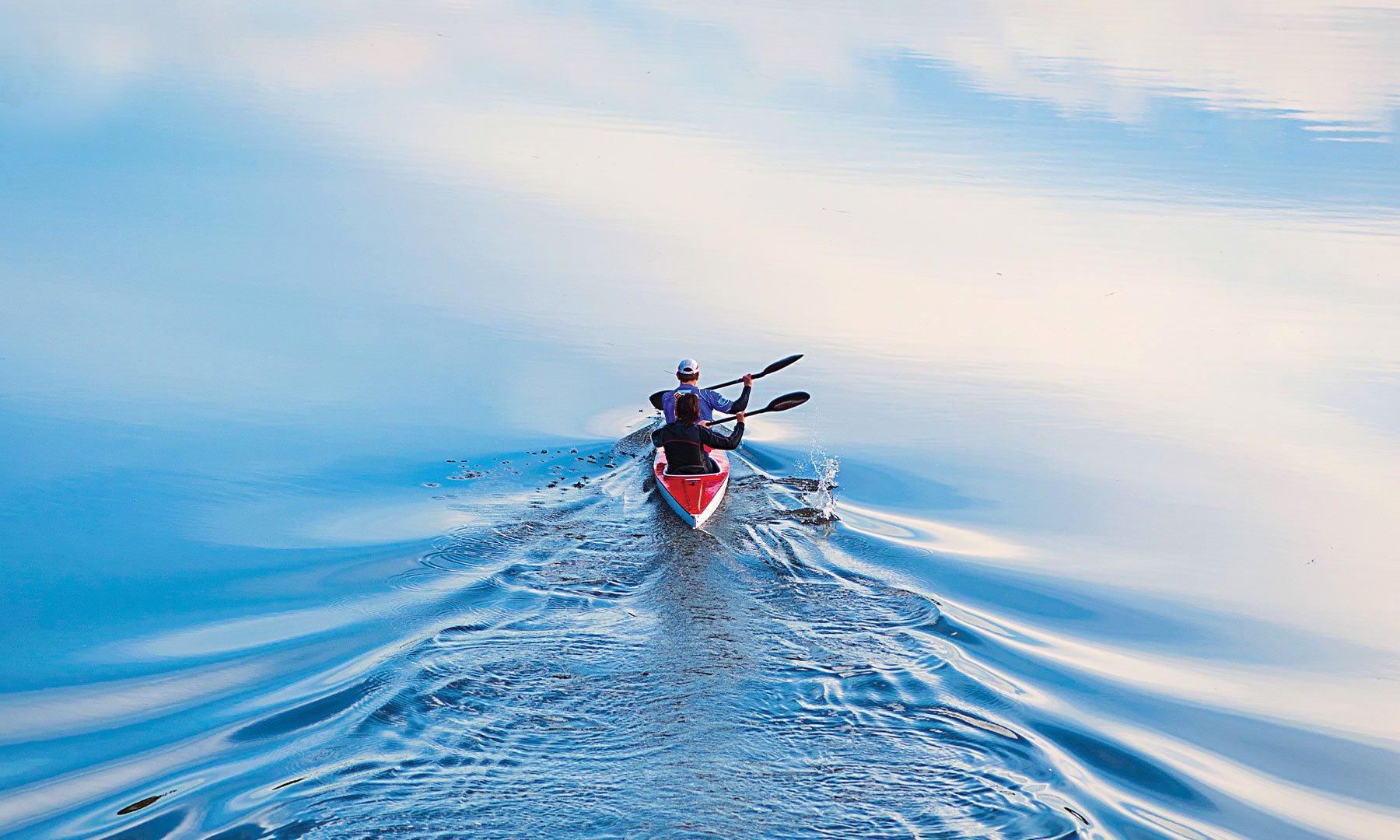 Image of couple rowing in red kayak on still blue water sky reflecting with ripples from kayak 